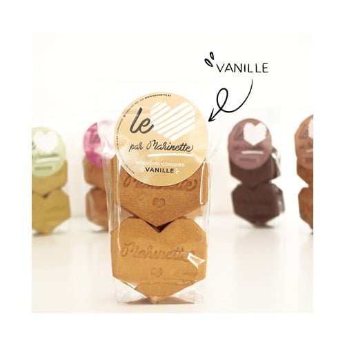 Un emballage en plastique transparent étiqueté « Cœurs de vanille » contient trois biscuits à la vanille en forme de cœur, chacun étant un biscuit personnalisé magnifiquement conçu. L'emballage est au premier plan, avec des emballages flous d'autres saveurs de biscuits visibles en arrière-plan. Une flèche pointe vers un petit texte « VANILLE » sur l'étiquette avant, soulignant sa saveur délicate.