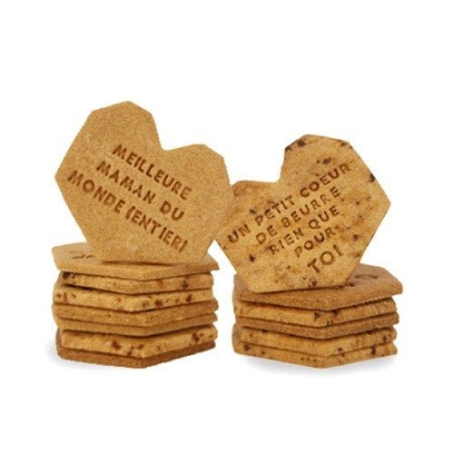 Deux piles de biscuits en forme de coeur avec inscriptions en français. Une pile indique « Meilleure maman du monde entier ! » (Meilleure maman du monde entier !), célébrant chaque *maman adorée* bien-aimée. L'autre indique "Un petit cœur du beurre, rien que pour toi". Chaque **biscuit personnalisé** ajoute une touche spéciale à ces messages sincères, les rendant parfaits pour toute occasion.