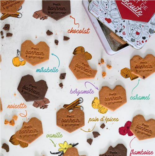 Un assortiment de petits bonheur à grignoter en forme de cœur aux saveurs de chocolat, de caramel, de vanille et bien plus encore est réparti autour d'une boîte à biscuits. Un biscuit personnalisé joliment décoré ajoute une touche spéciale à l'assortiment. Des illustrations d'ingrédients correspondant à chaque saveur sont dessinées à côté des friandises.